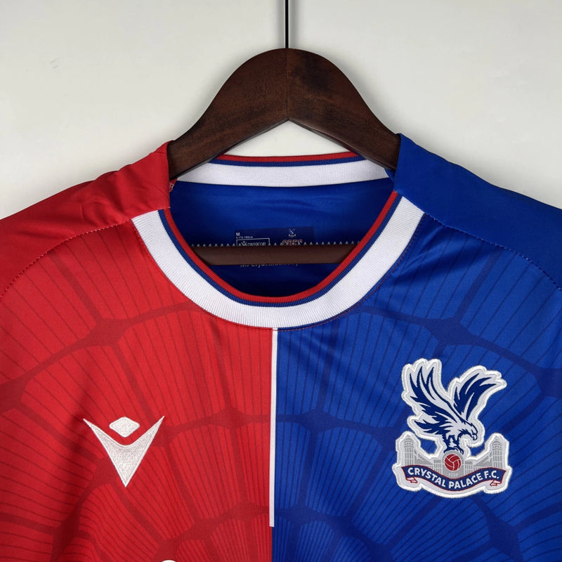 Camisa Crystal Palace Home I 2023/24 Torcedor Masculina - modelo azul com faixas verticais vermelhas, escudo do Crystal Palace bordado no peito e design tradicional da equipe londrina, confeccionada pela Macron.