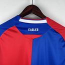 Camisa Crystal Palace Home I 2023/24 Torcedor Masculina - modelo azul com faixas verticais vermelhas, escudo do Crystal Palace bordado no peito e design tradicional da equipe londrina, confeccionada pela Macron.