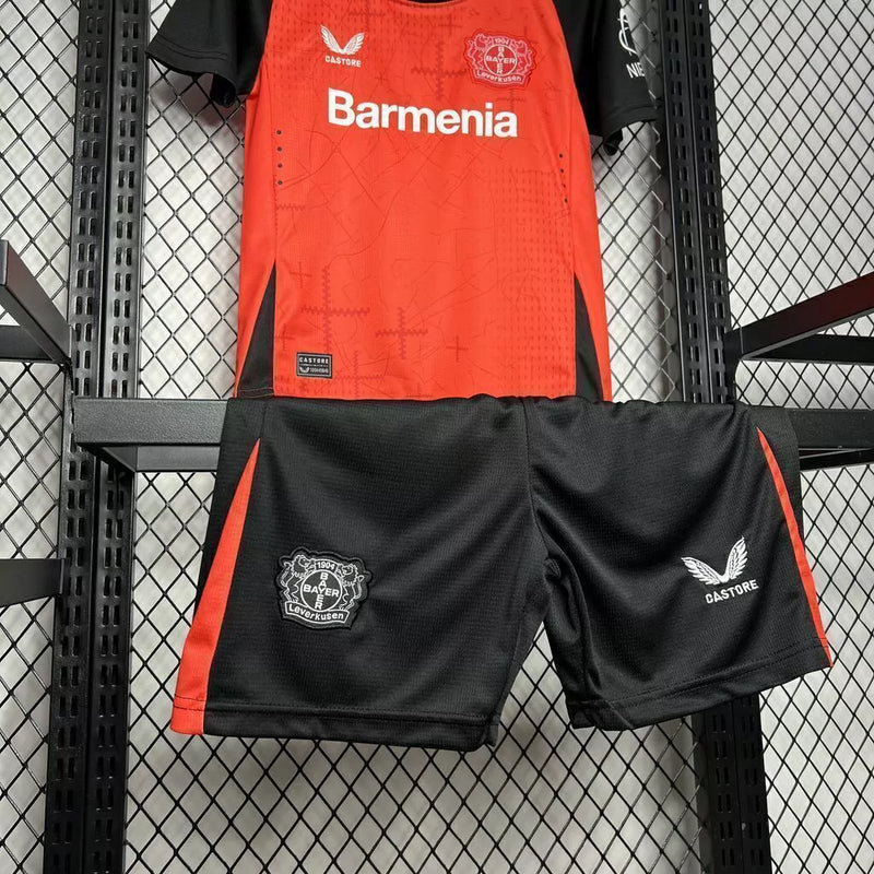 Kit Infantil Bayer Leverkusen Home 2024/25 Castore – uniforme oficial vermelho com detalhes pretos, composto por camisa e short, disponível do tamanho 3-4 anos ao 12-13 anos."