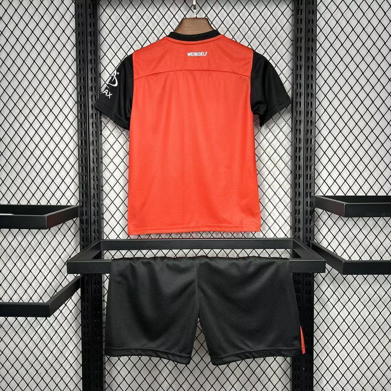Kit Infantil Bayer Leverkusen Home 2024/25 Castore – uniforme oficial vermelho com detalhes pretos, composto por camisa e short, disponível do tamanho 3-4 anos ao 12-13 anos."
