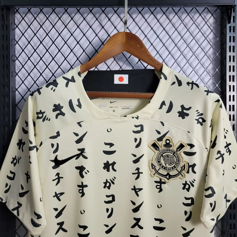 Camisa do Corinthians com estampa japonesa em katakana, escudo bordado no peito, modelo unissex em tom bege, pendurada em cabide sobre fundo de grade metálica. Design exclusivo estilo retrô edição limitada 2024.