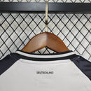 "Kit Infantil Alemanha Home 2024/25 Adidas – camisa branca com detalhes em preto e vermelho e shorts combinando, uniforme infantil oficial da seleção alemã, leve, respirável e confortável para crianças de 3 a 13 anos."
