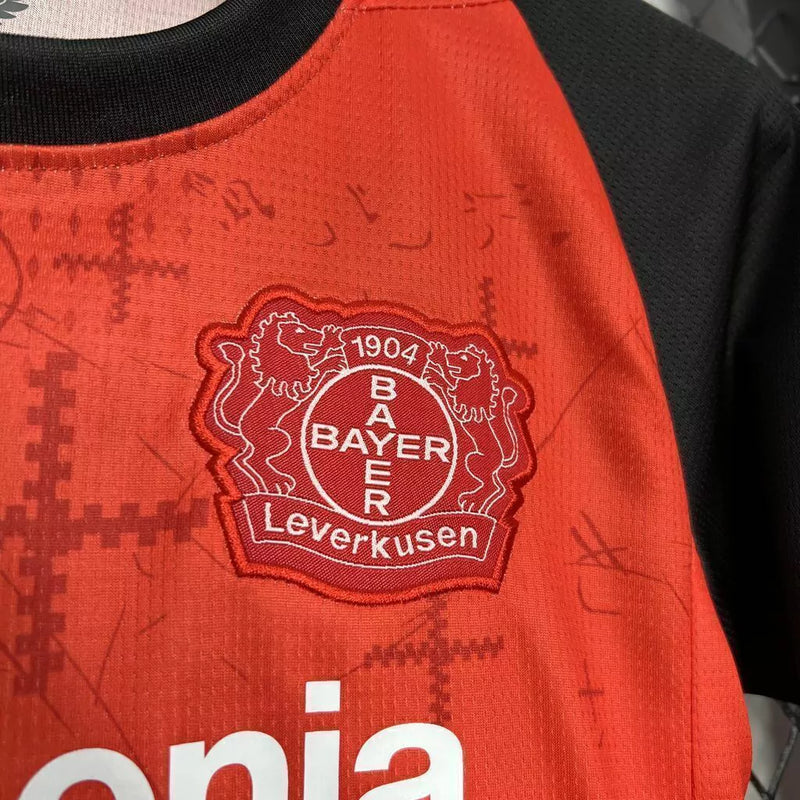 Kit Infantil Bayer Leverkusen Home 2024/25 Castore – uniforme oficial vermelho com detalhes pretos, composto por camisa e short, disponível do tamanho 3-4 anos ao 12-13 anos."