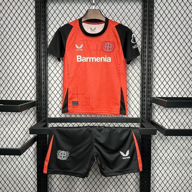 Kit Infantil Bayer Leverkusen Home 2024/25 Castore – uniforme oficial vermelho com detalhes pretos, composto por camisa e short, disponível do tamanho 3-4 anos ao 12-13 anos."