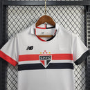 Kit Infantil São Paulo Home (1) 2024/25 New Balance oficial – camisa branca com listras vermelha, preta e branca, short e meião. Uniforme infantil do São Paulo FC temporada 2024/2025."
