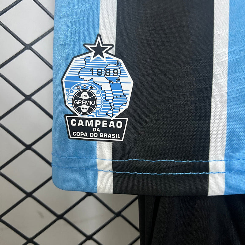 "Kit Infantil Grêmio Home 2024/25 Umbro, camisa azul, preta e branca listrada com shorts e meias combinando, uniforme oficial para crianças de 3 a 13 anos torcedoras do Grêmio."
