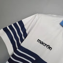 Camisa Lazio Home I 2014/15 Retrô Masculina branca com detalhes em azul celeste, gola em “V” azul e escudo bordado, representando a elegância e tradição do futebol italiano e da torcida Biancoceleste.