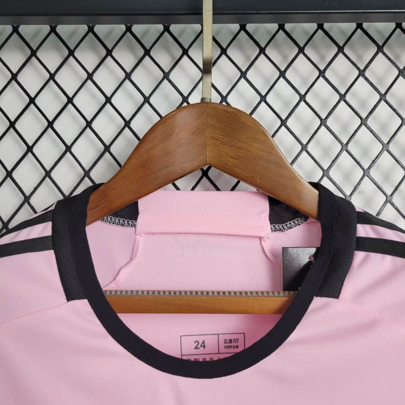 "Kit Infantil Inter Miami Home (1) 2024/25 Adidas oficial, camisa branca com detalhes em rosa e preto, escudo do Inter Miami CF em destaque, shorts e meias combinando, uniforme completo para crianças de 3 a 13 anos"