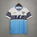 Camisa Lazio Home I 2014/15 Retrô Masculina branca com detalhes em azul celeste, gola em “V” azul e escudo bordado, representando a elegância e tradição do futebol italiano e da torcida Biancoceleste.