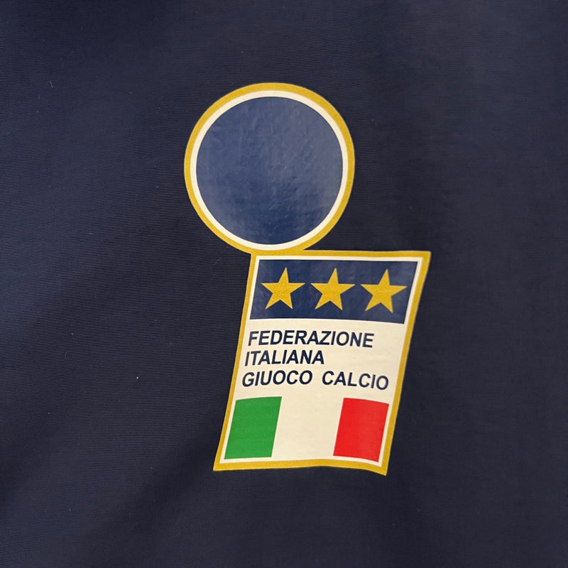 Corta-Vento Itália 2024/25 Adidas Masculino, azul, com escudo da seleção italiana e logo Adidas em destaque, tecido leve, respirável e resistente ao vento, com bolsos laterais e ajuste confortável, ideal para uso casual ou esportivo.”