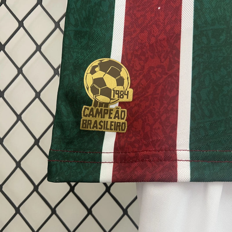 "Kit Infantil Fluminense Home 2024/25 Umbro completo para pequenos torcedores, com camisa tricolor verde, grená e branco bordada com escudo oficial e short correspondente, ideal para treinos, jogos e uso casual."