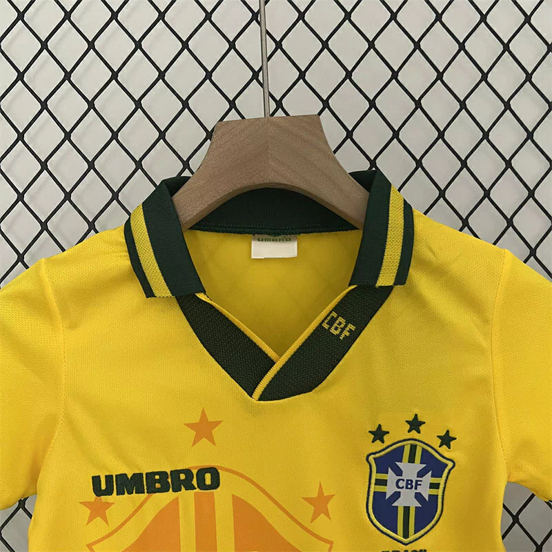 Kit Infantil Brasil Home 1994 Umbro, camisa amarela com detalhes verdes e azuis, shorts e meias combinando, uniforme retrô oficial para crianças de 3 a 13 anos torcedoras da Seleção Brasileira."