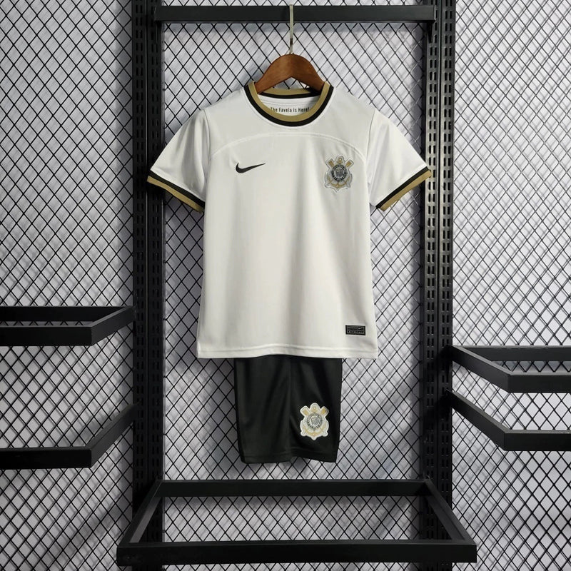 Kit Infantil Corinthians Home 2022/23 Nike, camisa branca com shorts e meias pretas, uniforme oficial para crianças de 3 a 13 anos torcedoras do Timão."
