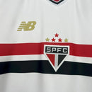 Kit Infantil São Paulo Home 2025/26 New Balance com camisa branca e faixas vermelha e preta, escudo do SPFC bordado, shorts combinando, modelo torcedor infantil, ideal para jogos, treinos e uso casual, oficial e autêntico."